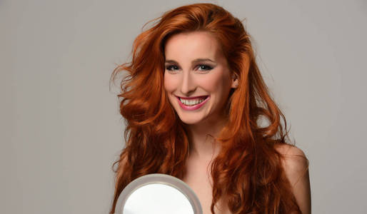 美丽的年轻女子与优雅的长红色闪亮的头发拿着镜子