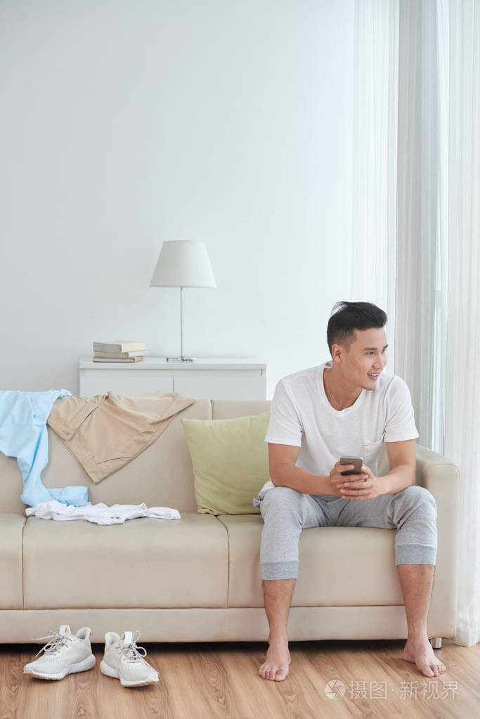 微笑着的亚洲年轻人坐在沙发上, 手里拿着智能手机