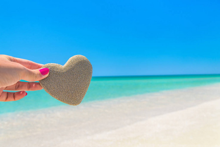心脏由沙子在女人手反对热带海洋海滩背景。浪漫, 爱情, 蜜月, 婚礼或圣情人节贺卡概念