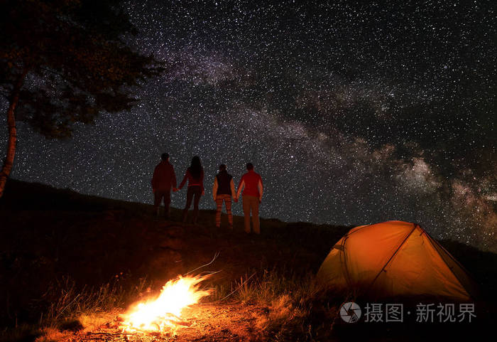 两对徒步旅行者牵手, 享受银河在星空下, 在一个篝火和橙色帐篷在山上