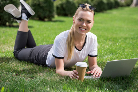 美丽的金发女孩微笑着在相机, 而躺在草地上, 使用笔记本电脑