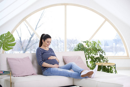 美丽的孕妇肖像坐在沙发上抚摸她的腹部, 而在家里放松
