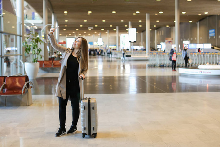 白人年轻女子去机场大厅与手提箱, 并使自拍由智能手机
