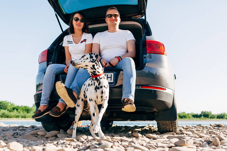 幸福的年轻夫妇与狗在 roadtrip 的肖像。男人和女人坐在格子上户外野餐概念