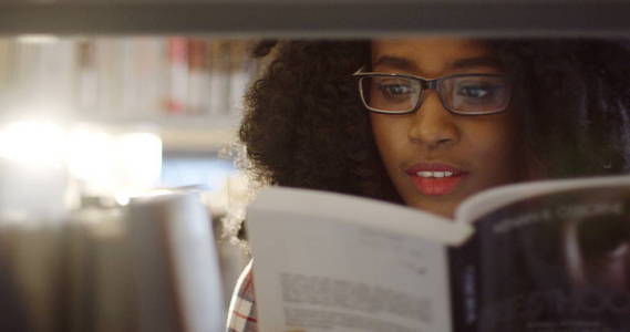 关闭的非洲裔美国年轻漂亮卷曲的女人通过图书馆书架上的书籍看。肖像拍摄。室内