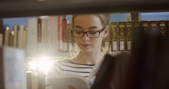 戴眼镜的年轻女学生的肖像照片在书架上看书, 书在图书馆里。关门了室内