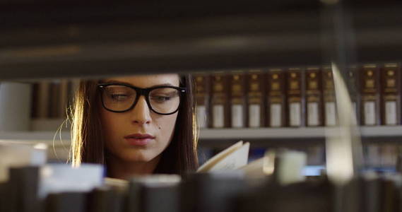 戴眼镜的年轻迷人的女学生在图书馆里书架上的书上翻书页。肖像.关门了内