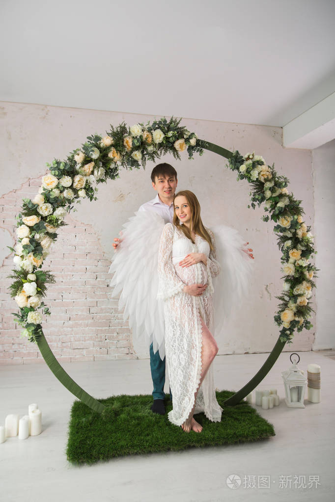 美丽的男人和怀孕的女孩在白色花边礼服下的装饰拱的花朵