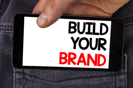 写笔记显示建立你的品牌。商业照片展示创建您自己的徽标口号模型广告电子营销写在手机屏幕上的人持有的牛仔裤背景