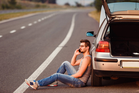 一个年轻人坐在柏油上, 靠在他的车上, 然后疏散.他的车抛锚了.照片