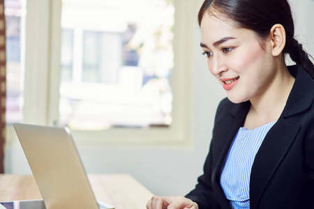穿着黑色西装的微笑的商业女性正坐在办公室里的笔记本电脑上工作。快乐工作理念