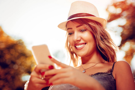 美丽的微笑年轻女子与夏天的帽子在大自然中享受秋天和使用智能手机