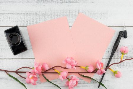 两张空白粉红色卡片, 斜笔和墨水瓶用假粉色花枝装饰在白色木头背景上