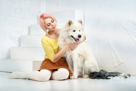 有吸引力的年轻女孩与短的金发在黄色毛衣和裙子拥抱她心爱的宠物狗养殖萨摩耶
