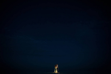 新娘和新郎站在沙漠里, 在夜晚的灯泡花环旁边牵手。