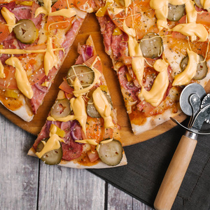 特写比萨与番茄酱, 奶酪酱, 意大利干酪, 西红柿, 香肠, 泡菜, 甜洋葱, 黄色的辣椒在木制比萨饼板上, 比萨饼刀和深色餐巾