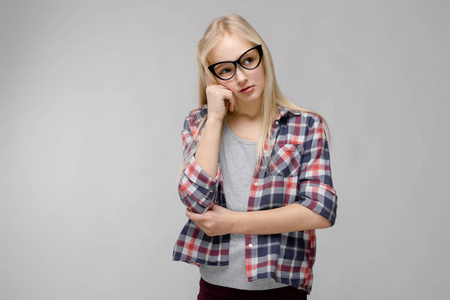漂亮的白种金发女孩在格子衬衫和眼镜显示不同的情感表现在灰色墙在演播室。用手在脸上思考