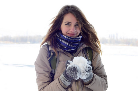 一个年轻而快乐的白种女孩穿着棕色大衣, 在冬天的天空和冰冻的湖泊的地平线背景下举行雪球。