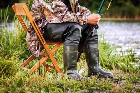 渔夫坐在军装和橡胶靴的椅子上, 在湖岸边钓鱼竿