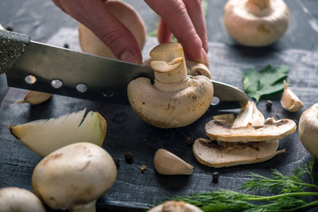 女人的手正在用刀在黑暗的厨房桌子上切 champignons。烹调蘑菇菜肴的概念。特写