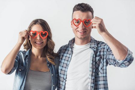 美丽的年轻夫妇在休闲服装持有两个红色的心形纸眼镜, 看着相机和微笑, 孤立的白色