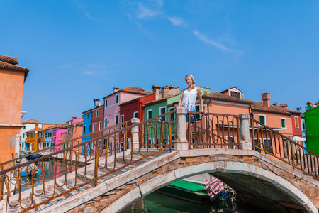 一个女游客站在一座横跨运河的桥上, 在布拉诺的许多五颜六色的房子背景