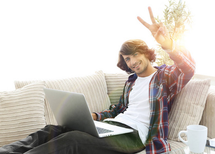 快乐的人，与笔记本电脑坐在沙发上，显示他的手一个胜利的手势