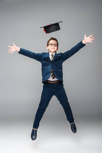 男孩的商人服装跳跃与毕业帽隔离灰色