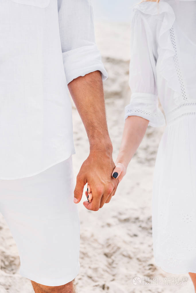 在沙丘前牵手的白色衣服中的一对夫妇的裁剪照片