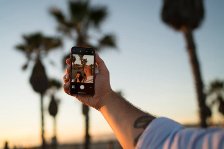 被裁剪的人的手持有现代智能手机, 使自拍他的照片与女友, 年轻男性使用前手机相机和应用程序拍摄视频在暑假旅行