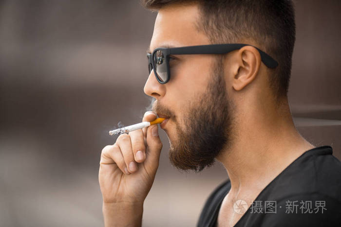 一个年轻英俊的白胡子男子在太阳镜和黑色 t恤烟在街上吸烟的春天.