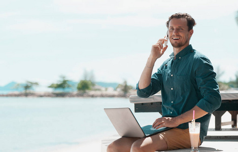 男子自由职业者与笔记本电脑和手机坐在沙滩上