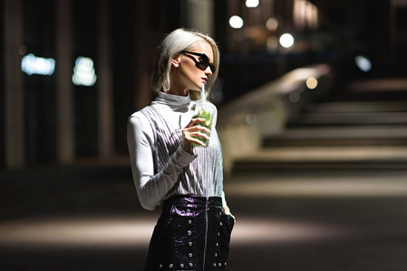 有吸引力的年轻妇女与莫吉托在塑料杯子在黑暗的街道