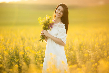 美丽的妇女与野花花束在黄色领域在日落灯, 夏天时间