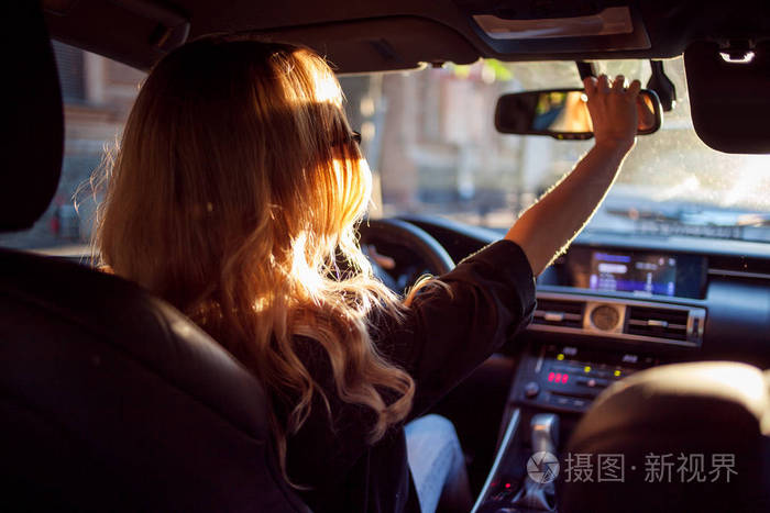 戴着太阳镜的漂亮年轻女子坐在车的车轮后面, 校正镜子.照片从后座