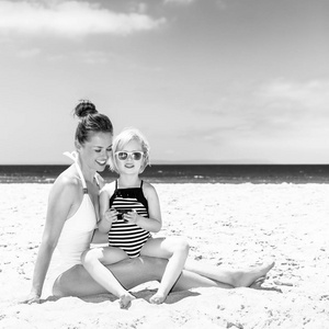 在白色沙滩上的家庭乐趣。快乐的现代母亲和女儿在海边的泳衣上观看照片在相机上