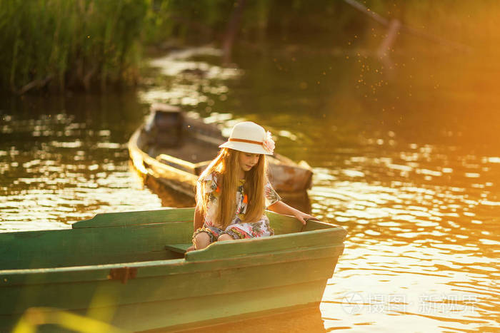 可爱的小女孩在一顶帽子坐在船上的湖泊日落