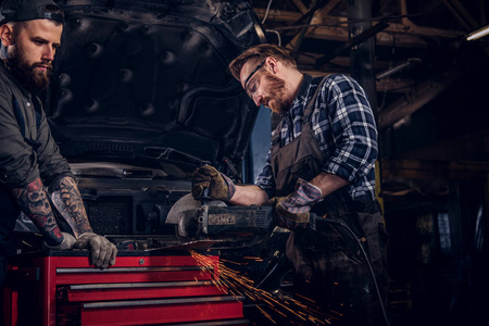 两个胡子的汽车技工在一个统一的和安全的眼镜工作的角度磨床, 而站在一个破碎的汽车在修理车库