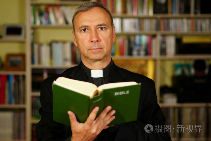 一个看起来很严肃的天主教神父正在学习, 读圣经, 进入他的图书馆.