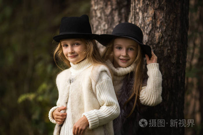 两个快乐的女孩同样穿着在森林的毛皮背心和帽子公园里的小女朋友儿童