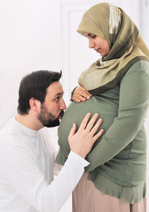 怀孕的穆斯林妻子与丈夫亲吻的肚子和他的手在肚子上微笑
