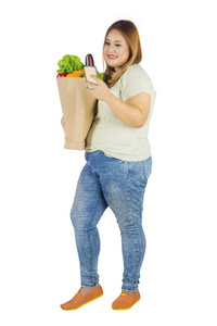 全长的白种人脂肪妇女手持一袋蔬菜, 而使用智能手机, 孤立的白色背景