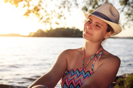 在一个温暖的夏日傍晚, 戴着帽子的漂亮女孩坐在湖岸边。室外城市外的年轻女子