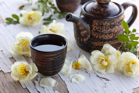 陶瓷杯, 茶壶和白狗玫瑰在花园里的旧木桌上。香薰茶与野玫瑰