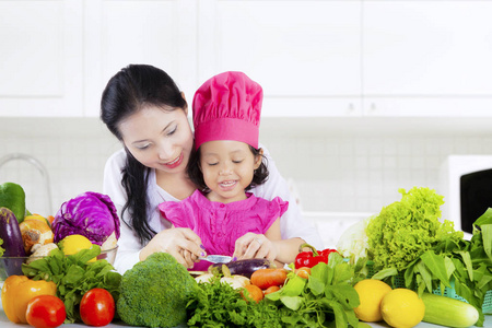 漂亮的年轻的亚洲妇女和她的小女儿在厨房的桌子上切蔬菜在家里