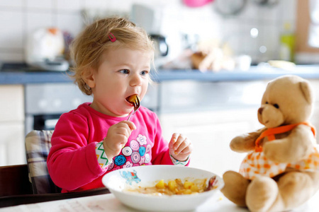 可爱的小女孩吃叉菜和面食。小孩子喂养和玩玩具泰迪熊