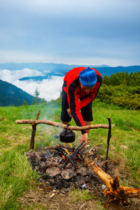 冒险家投掷木柴在篝火在一个绿色草甸反对山范围的背景在雾以后风暴。史诗般的旅行在山上