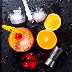 龙舌兰酒日出鸡尾酒, 橙色, 冰块, 樱桃樱桃, 冰钳和在湿黑色石板托盘上的跳汰机。鸡尾酒套装。平躺