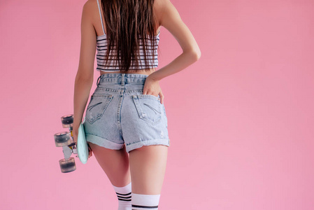 在粉红色背景下的年轻亚裔少女的裁剪图像。滑板女孩在短裤与滑板隔离