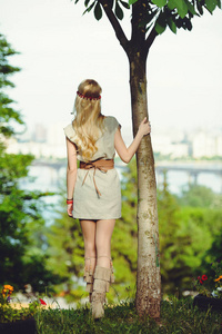 温柔的金发女郎抱着树干, 看着远处, 穿着自然的连衣裙, 时髦而舒适。自然的爱的概念